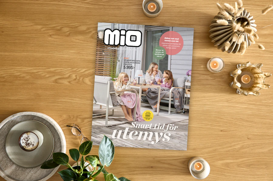 Kampanjblad för utemöbler hos Mio liger på ett skrivbord omgivet av ljuslyktor och växter. 