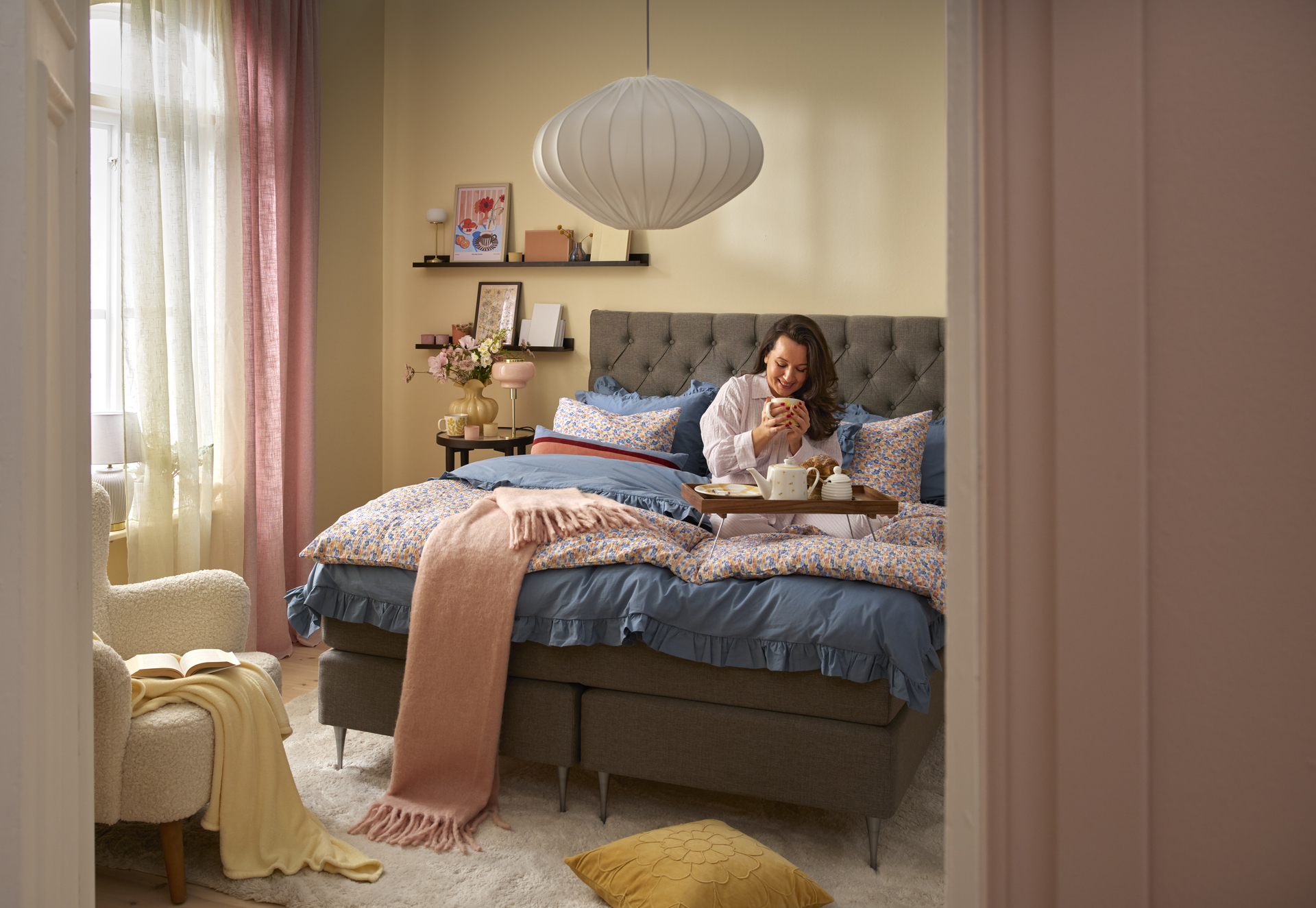 Kvinna sitter i kontinentalsäng i sovrumsmiljö med varma vårliga färgtoner. 