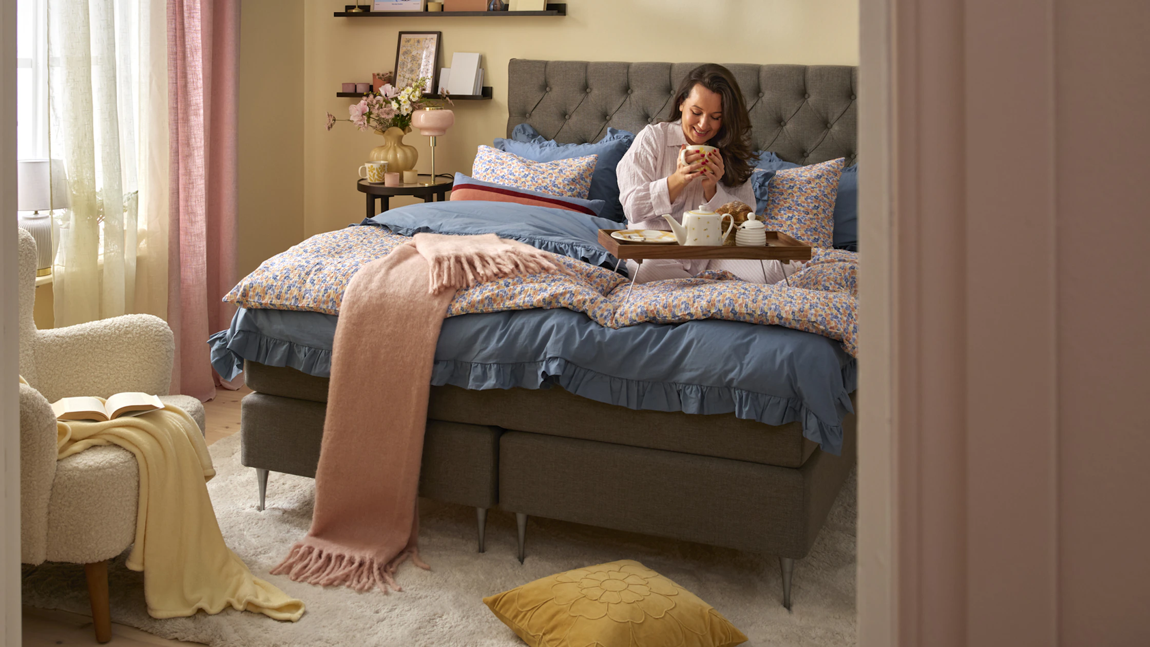 Kvinna sitter i kontinentalsäng i sovrumsmiljö med varma vårliga färgtoner.