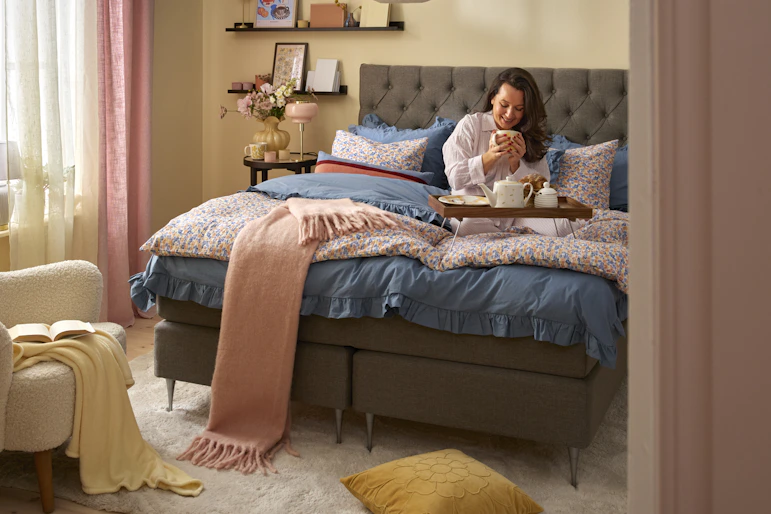 Kvinna sitter i kontinentalsäng i sovrumsmiljö med varma vårliga färgtoner.