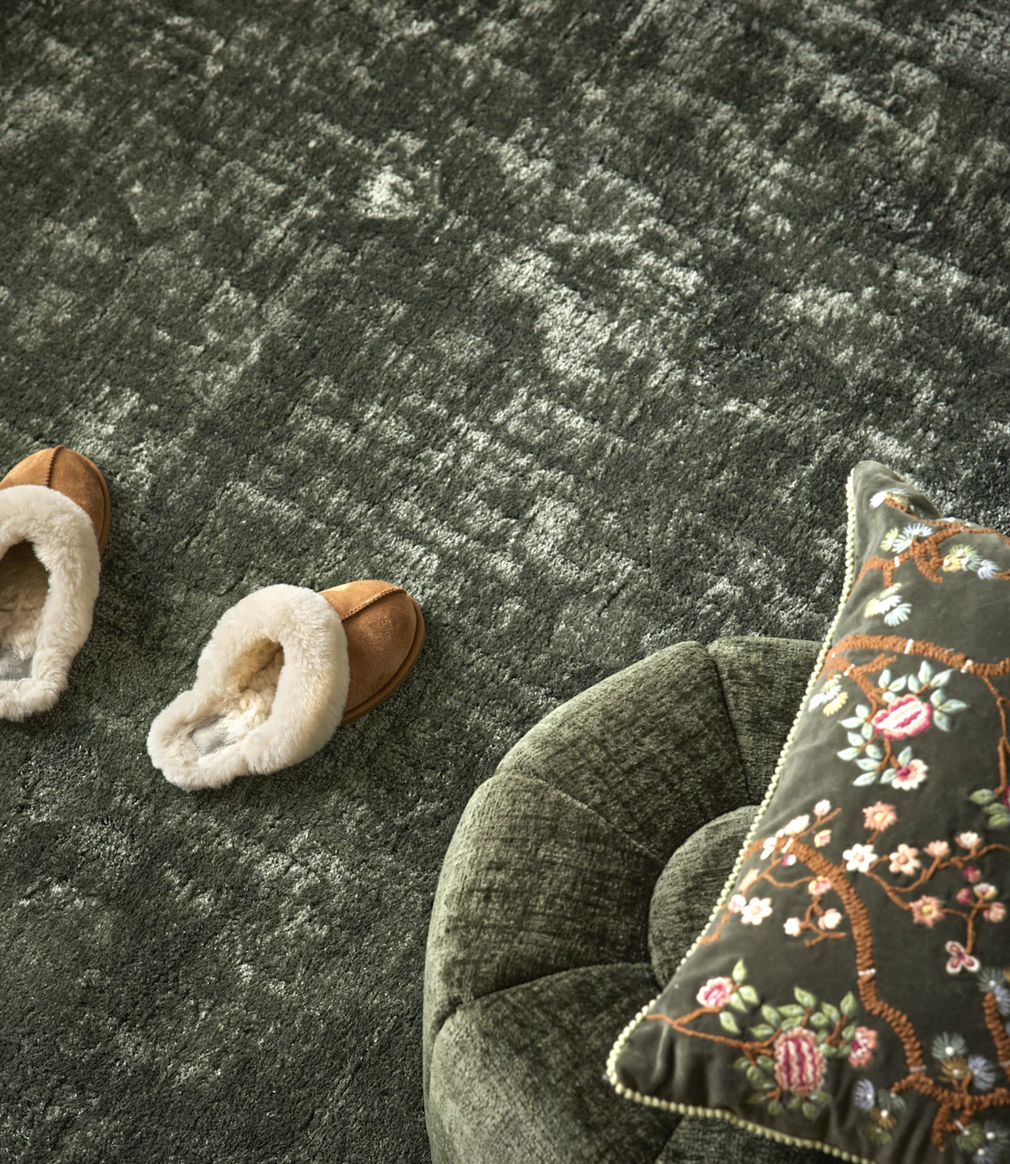 Grön matta på golv med fårskinnstofflor som står på, i förgrund syns del av en fotpall med mönstrad prydnadskudde.