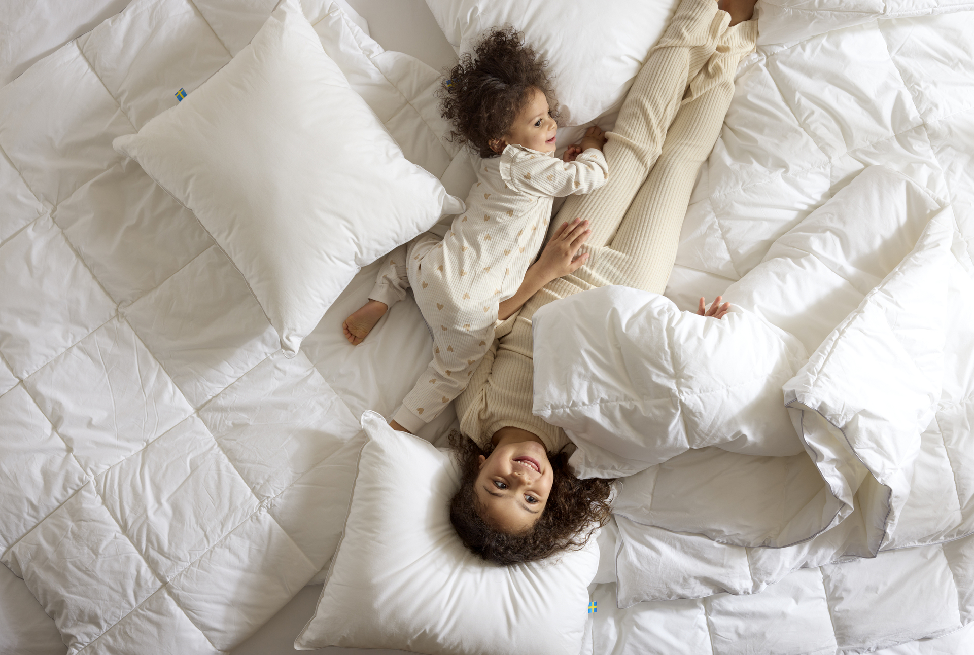 Två små barn busar bland täcken och kuddar i en obäddad säng.