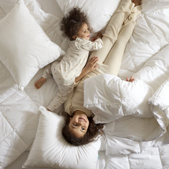 Två små barn busar bland täcken och kuddar i en obäddad säng.