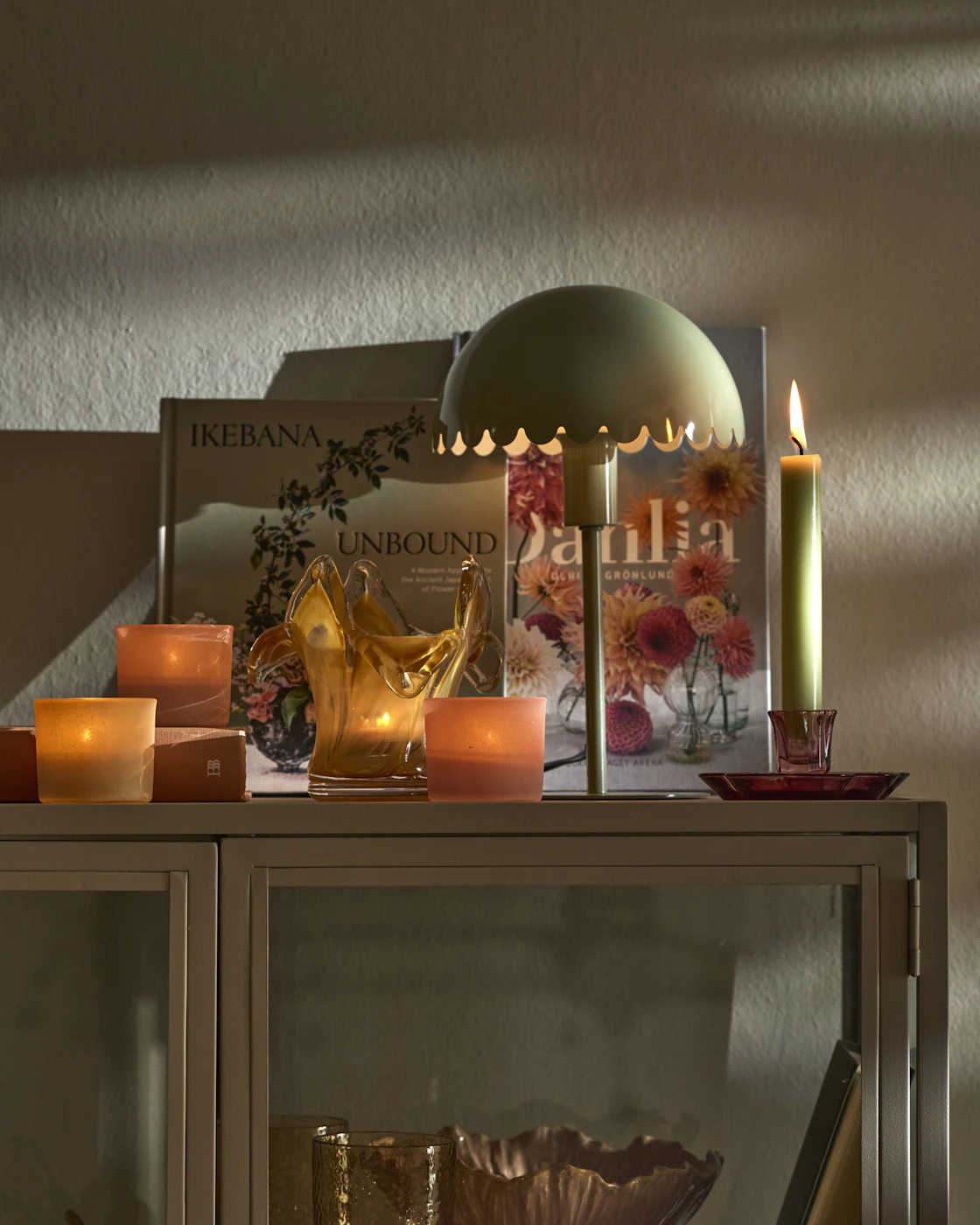 Flera ljuslyktor, en grön lampa och en ljusstake med ett grönt ljus står på ett vitrinskåp.