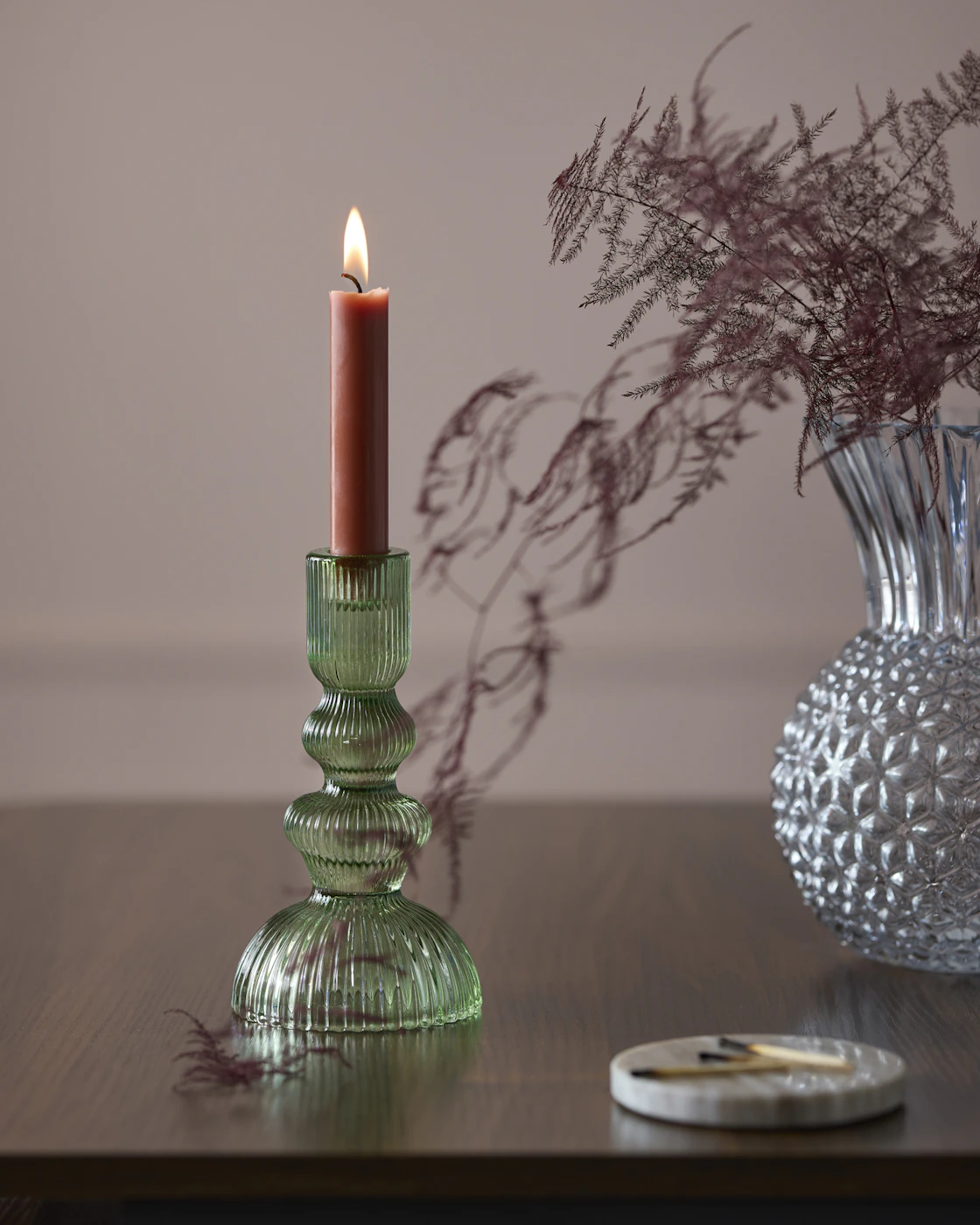 En grön ljusstake med ett rustikljus och en vas med en mörklila växt står på ett bord.