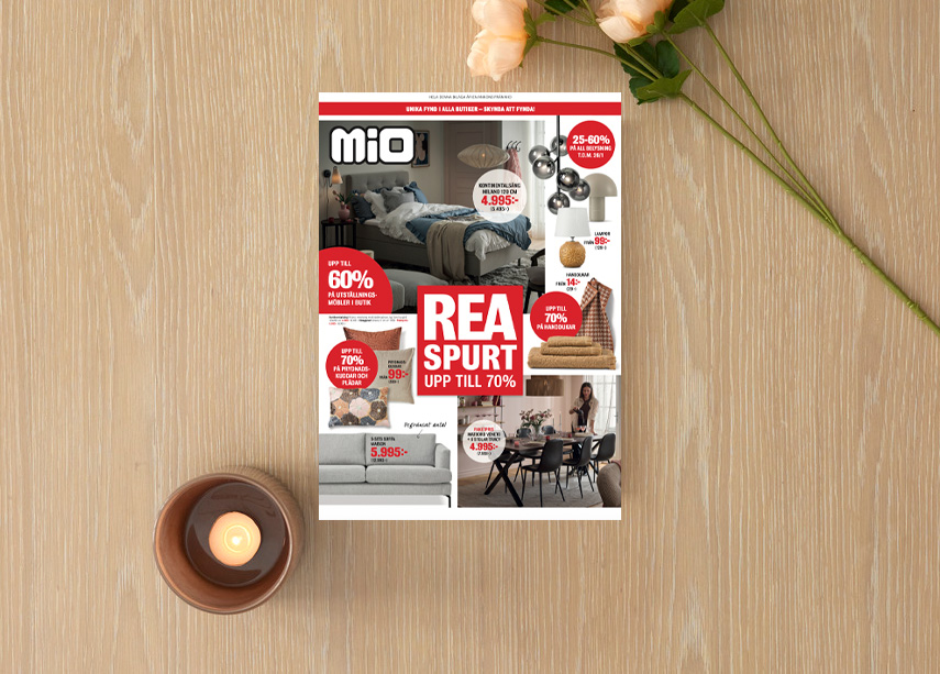 Ett kampanjblad för rea ligger på ett ljust träbord med en blomma och ljuslykta bredvid 