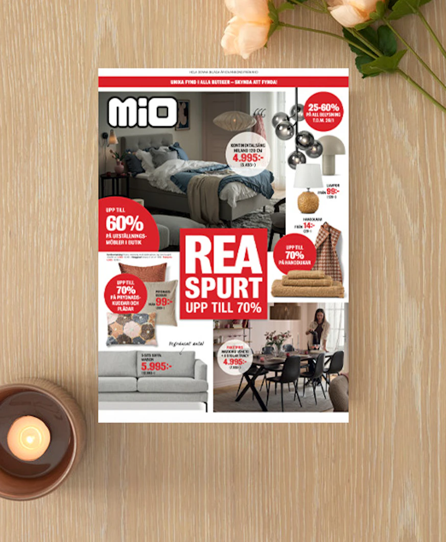 Ett kampanjblad för rea ligger på ett ljust träbord med en blomma och ljuslykta bredvid