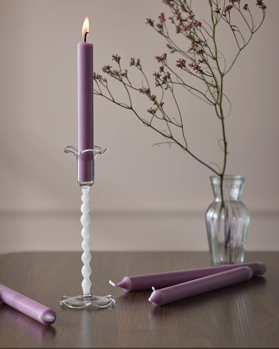 En swirlad vit ljusstake och ett lila rustikljus står framför en vas med en lila snittblomma.