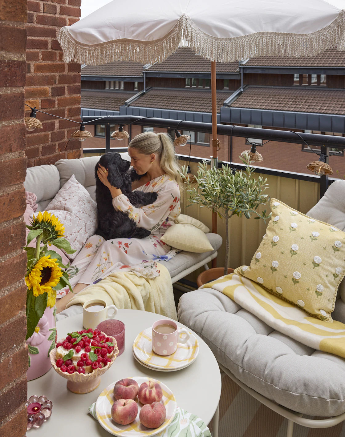 En beige utefåtölj och matchande utesoffa står på en balkong. I soffan sitter en kvinna och gosar med en svart hund.