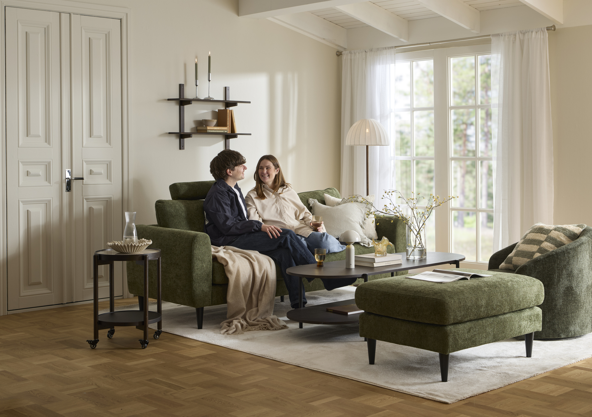 ett par sitter i en grön soffa placerad i ett ljust vardagsrum vid ett stort fönster