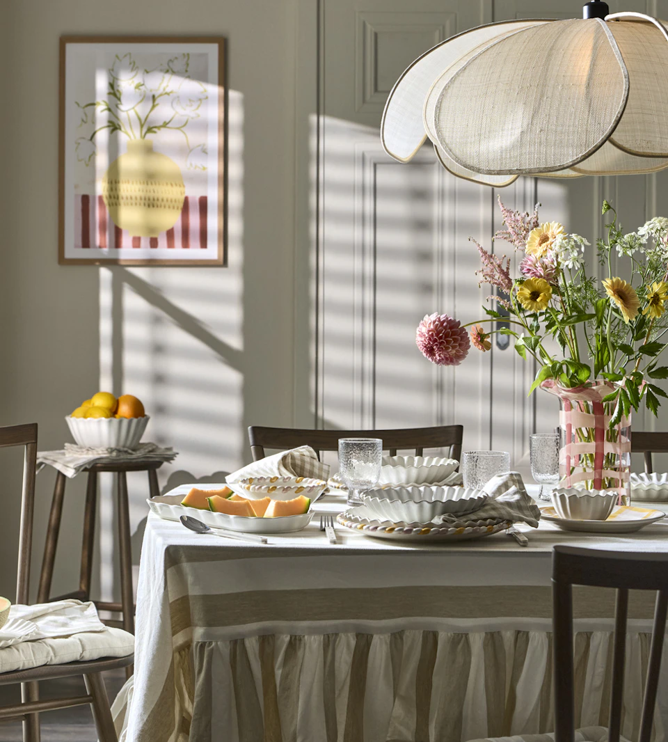 ett bprd med en randig duk är dukat med vitt porslin och en stor rosa vas med blommor