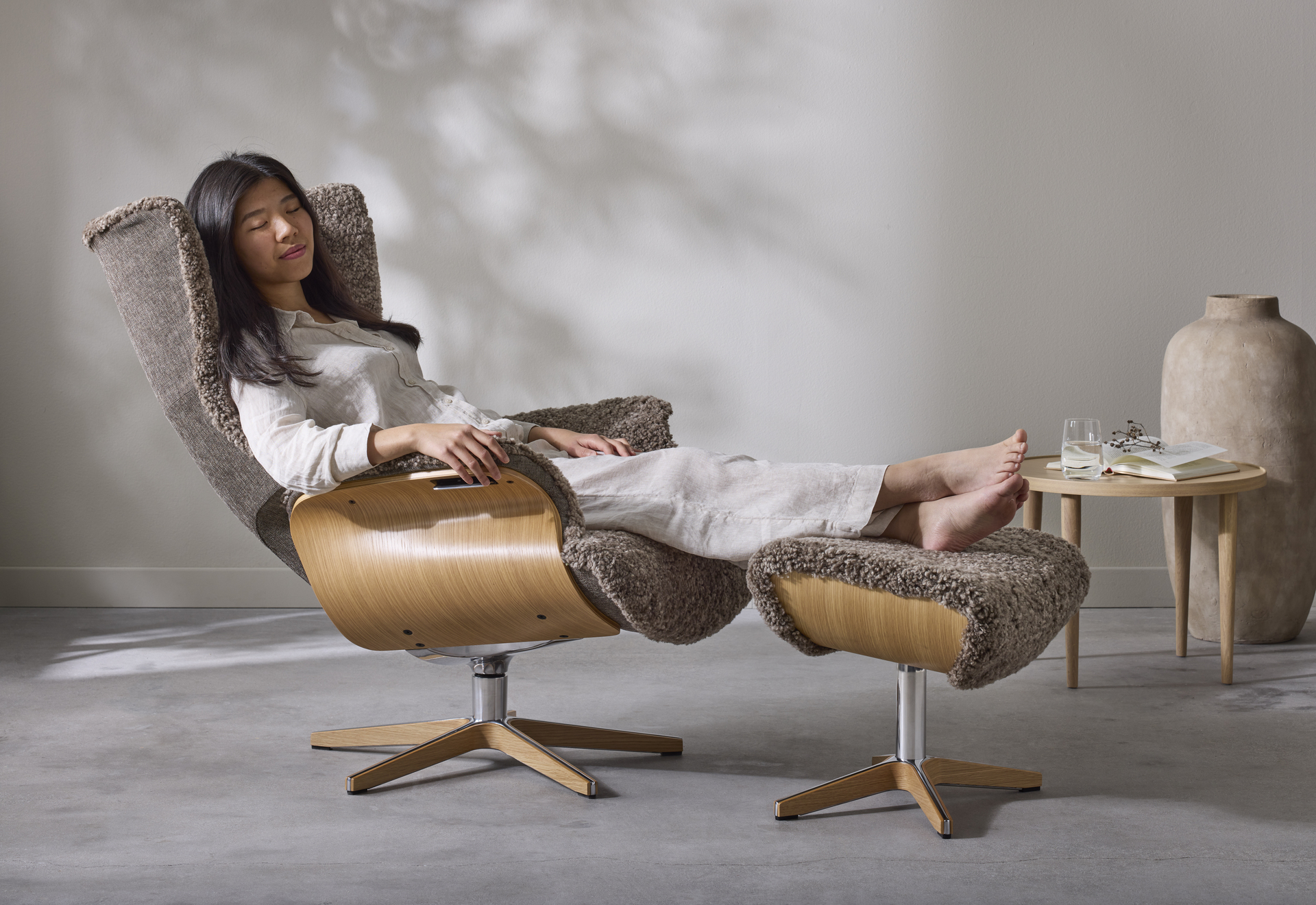 I en brun fårskinnsfåtölj med underrede i ek sitter en kvinna bakåtlutad med fötterna på en fotpall