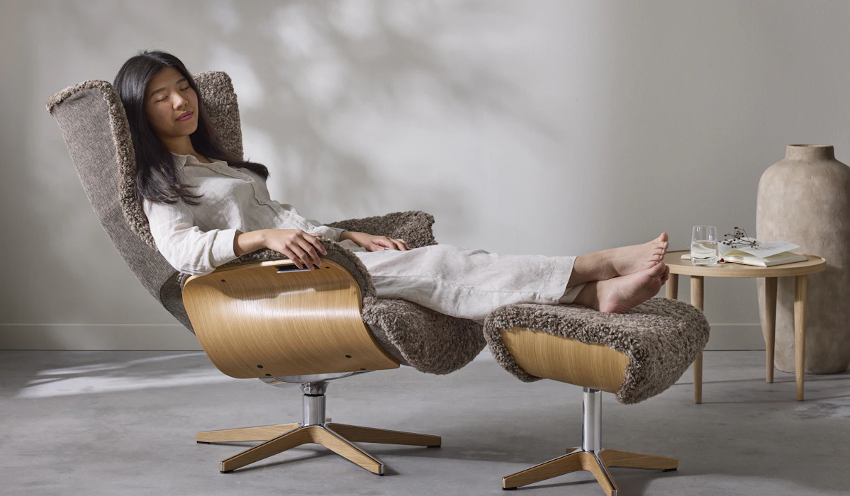 I en brun fårskinnsfåtölj med underrede i ek sitter en kvinna bakåtlutad med fötterna på en fotpall