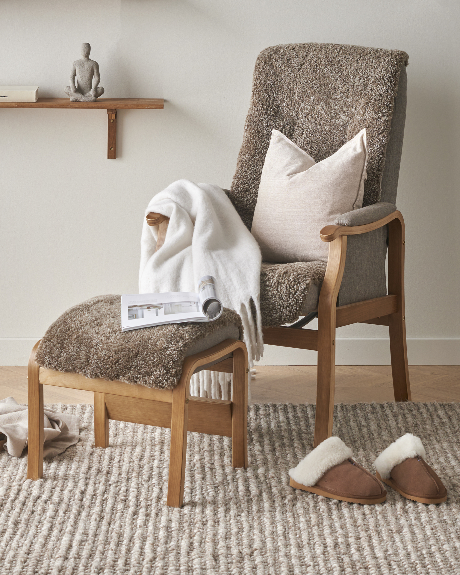I en brun fårskinnsfåtölj ligger en beige kudde och en vit filt