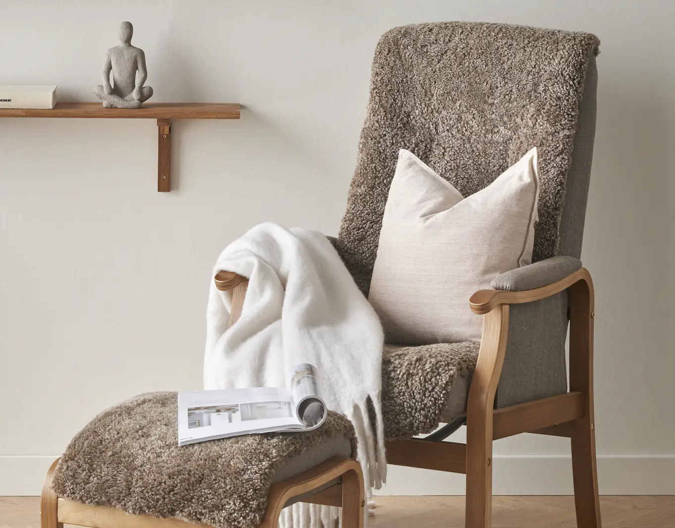 I en brun fårskinnsfåtölj ligger en beige kudde och en vit filt