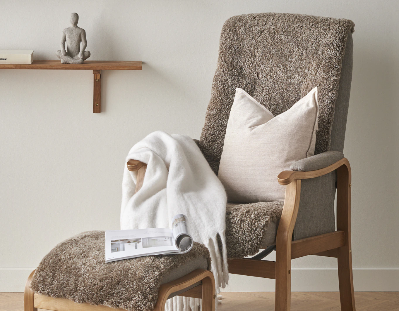 I en brun fårskinnsfåtölj ligger en beige kudde och en vit filt
