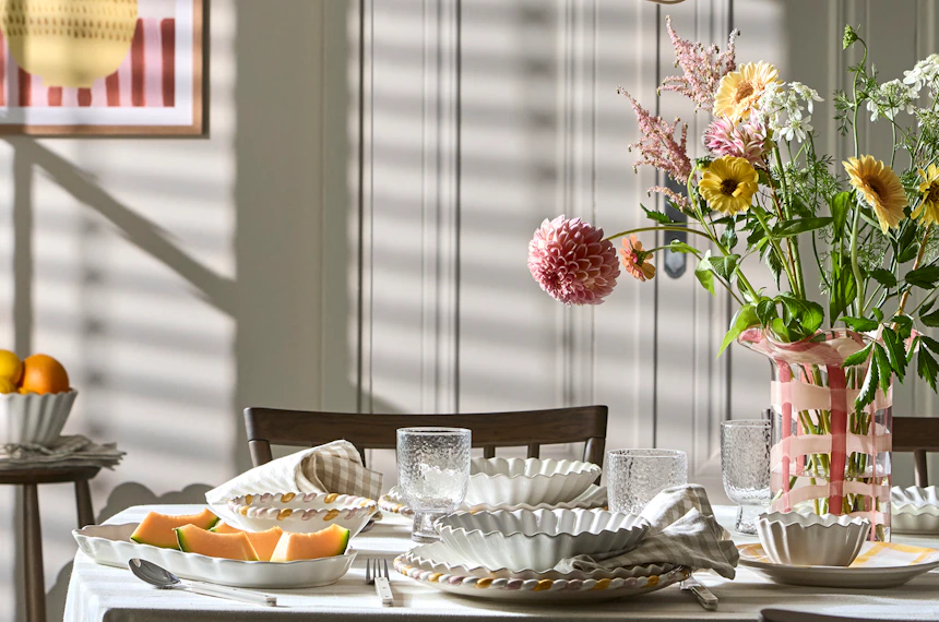 ett bord med en randig duk är dukat med vitt porslin, mat, och en rosa vas med blommor i