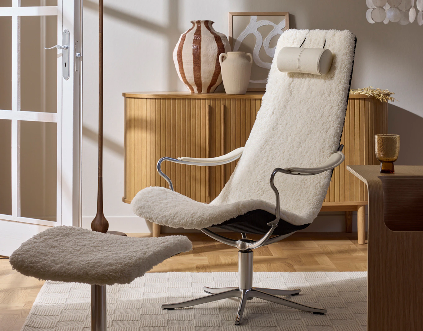 I ett beiget rum står en vit fåtölj i fårskinn framför en ekfärgad skänk