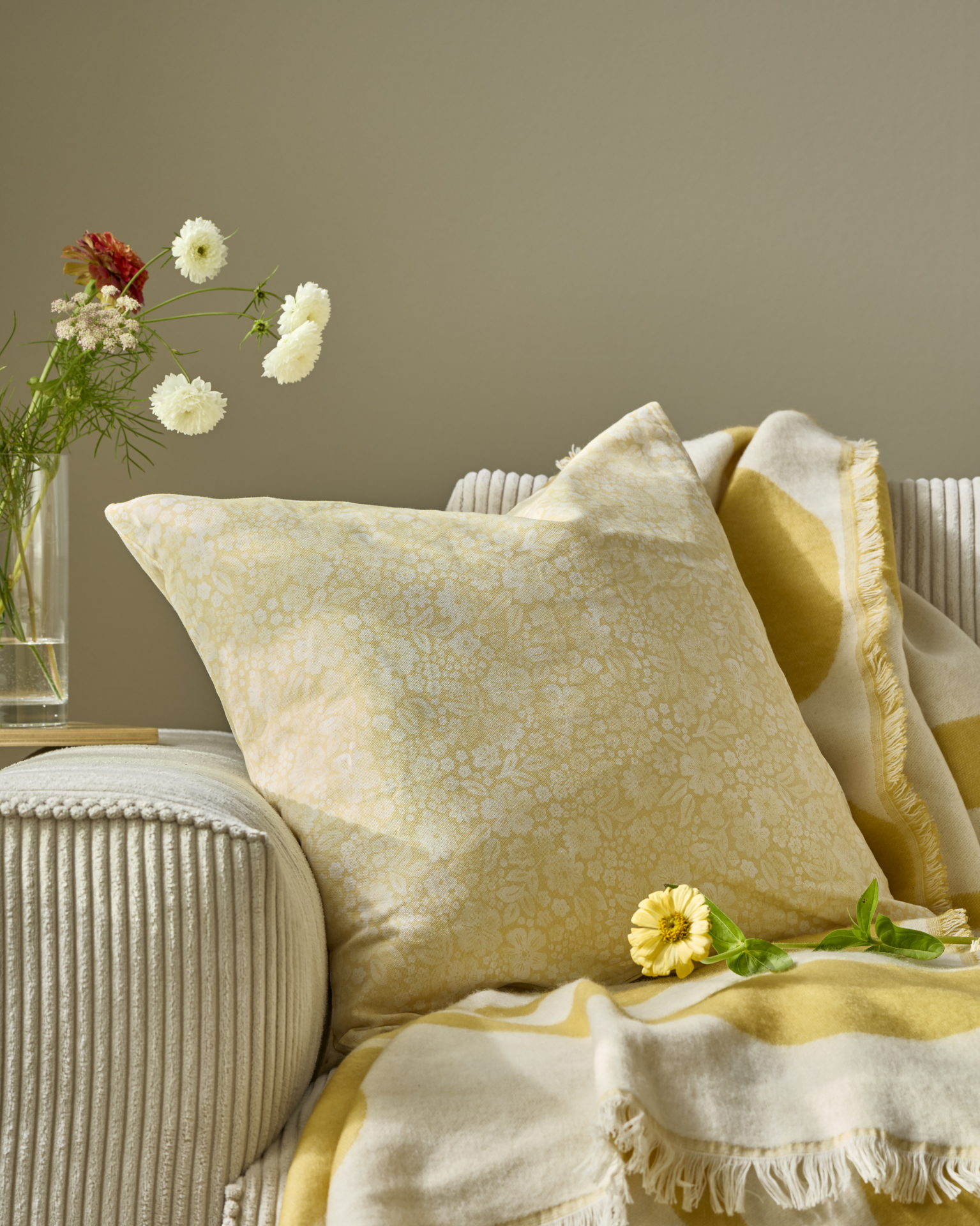 en gulmönstrad kudde ligger i en beige soffa med en gul pläd och en vas med blommor bredvid