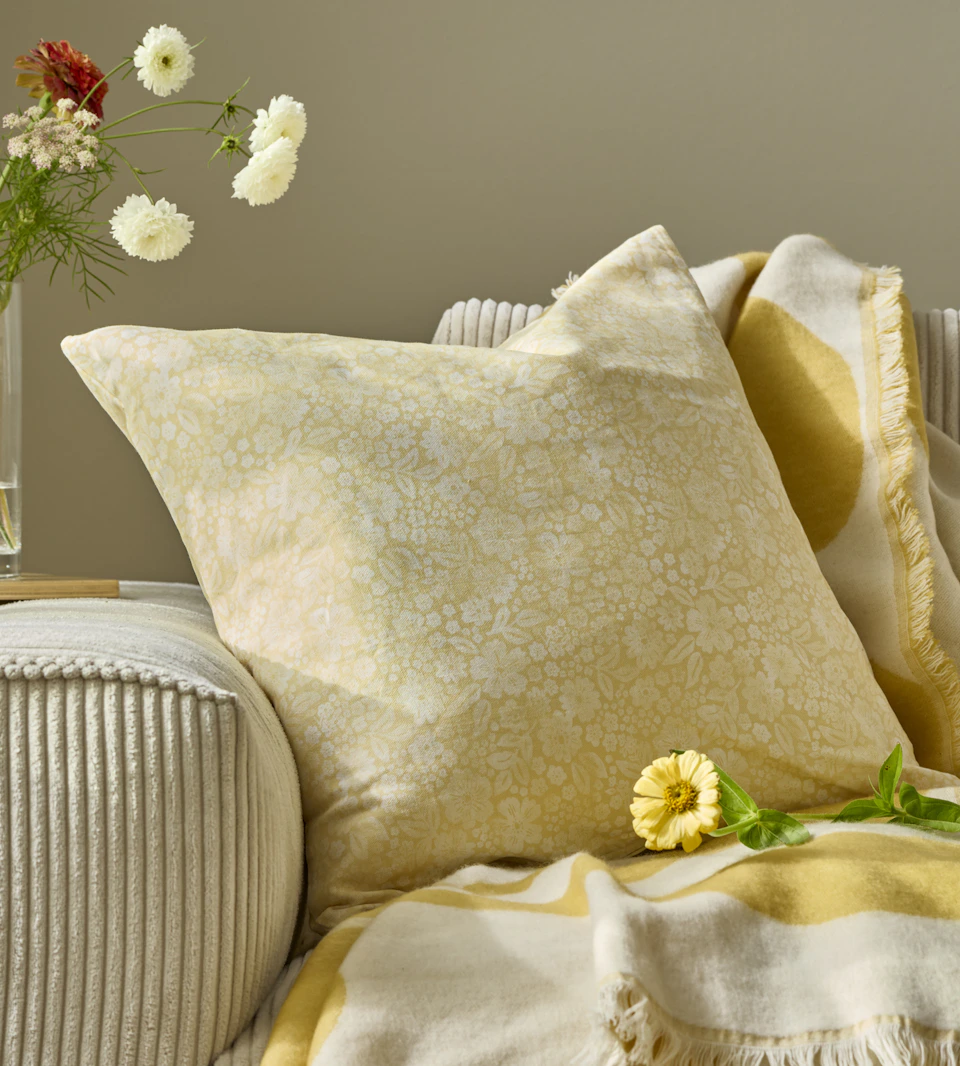 en gulmönstrad kudde ligger i en beige soffa med en gul pläd och en vas med blommor bredvid
