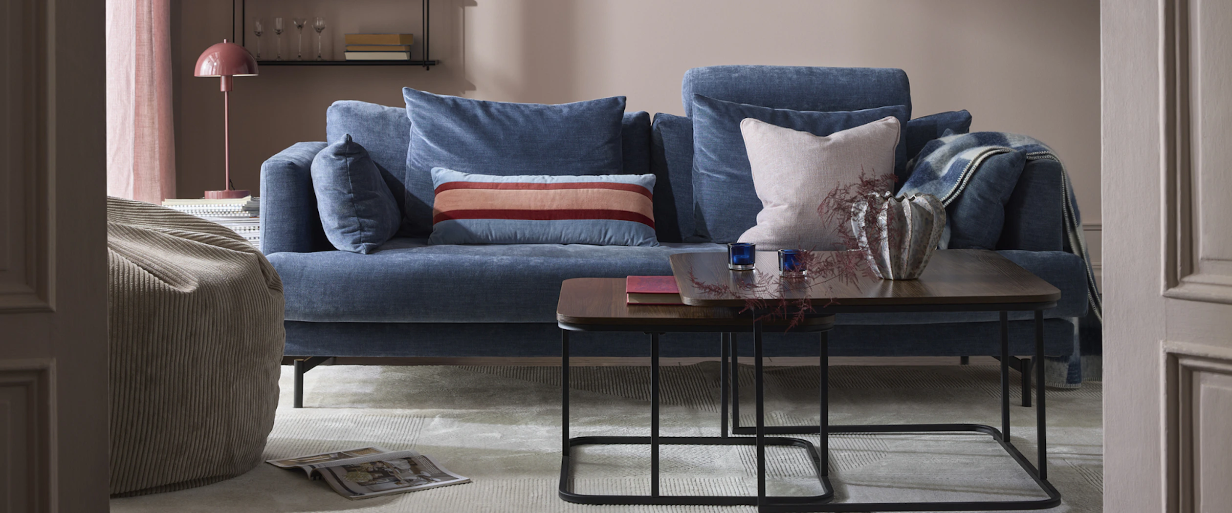 I ett rosa vardagsrum står en blå soffa i sammet bakom ett soffbord i mörkt trä