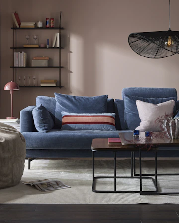 I ett rosa vardagsrum står en blå soffa i sammet bakom ett soffbord i mörkt trä