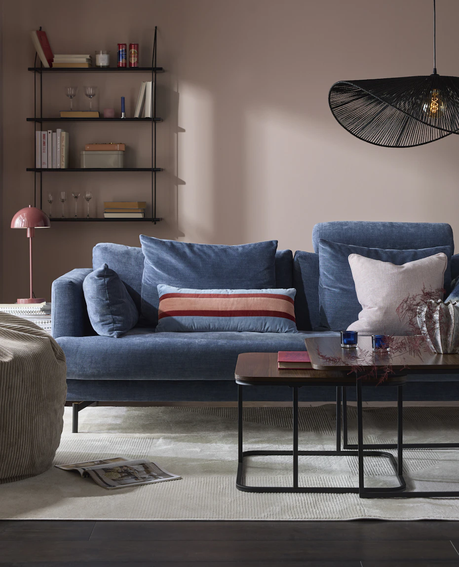 I ett rosa vardagsrum står en blå soffa i sammet bakom ett soffbord i mörkt trä