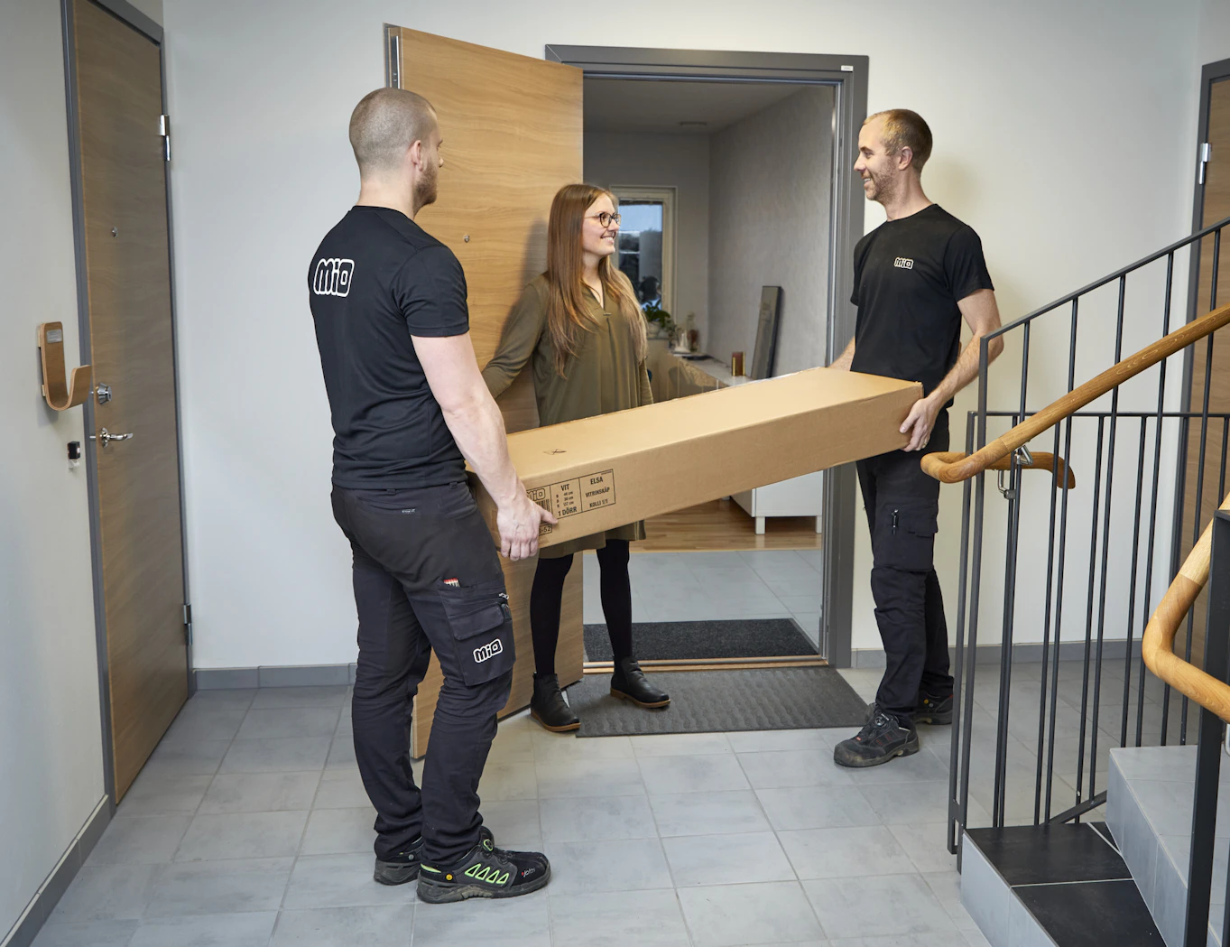 Trapphus där två män i svarta kläder levererar en stor låda till kvinna som håller upp dörren till sin lägenhet. 