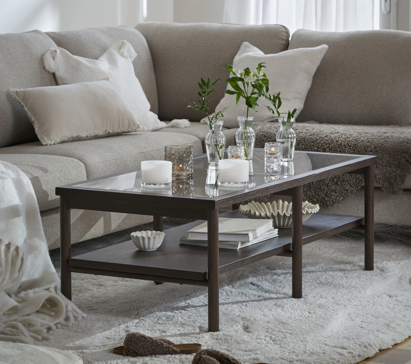 Framför en ljusgrå L-formad soffa står ett avlångt soffbord i mörkt trä och glas med en hylla under