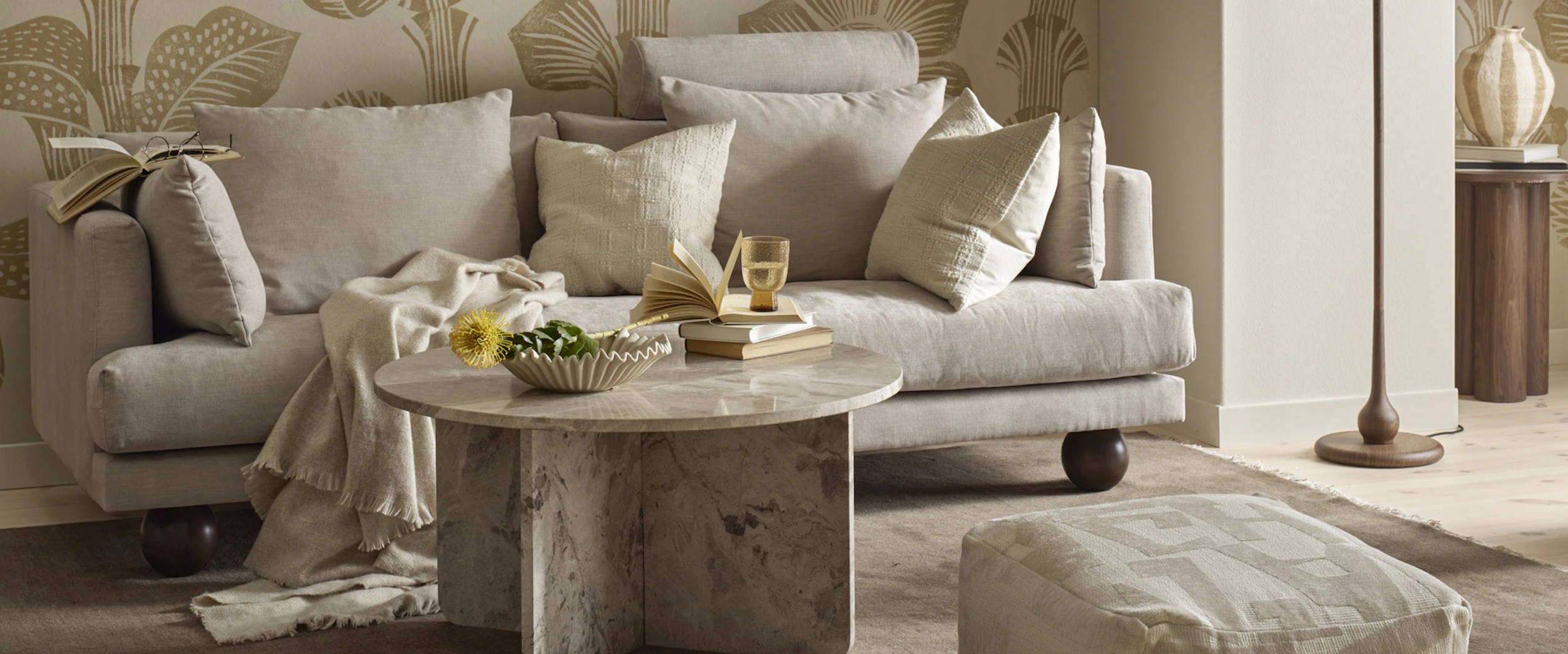 I ett vardagsrum står en beige soffa med klotformade ben bakom ett runt soffbord i beige marmor