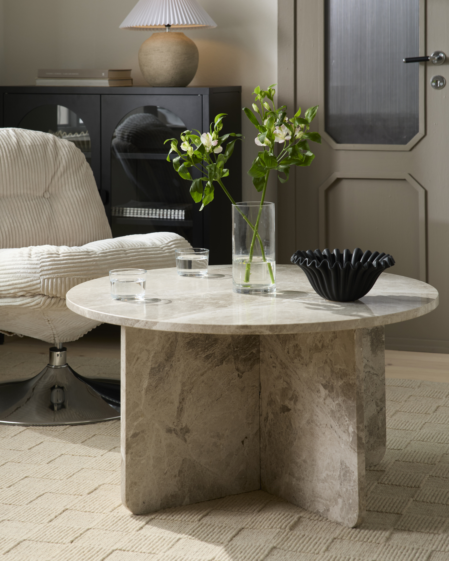 Ett runt soffbord i beige matmor står intill en fåtölj i beige manchester