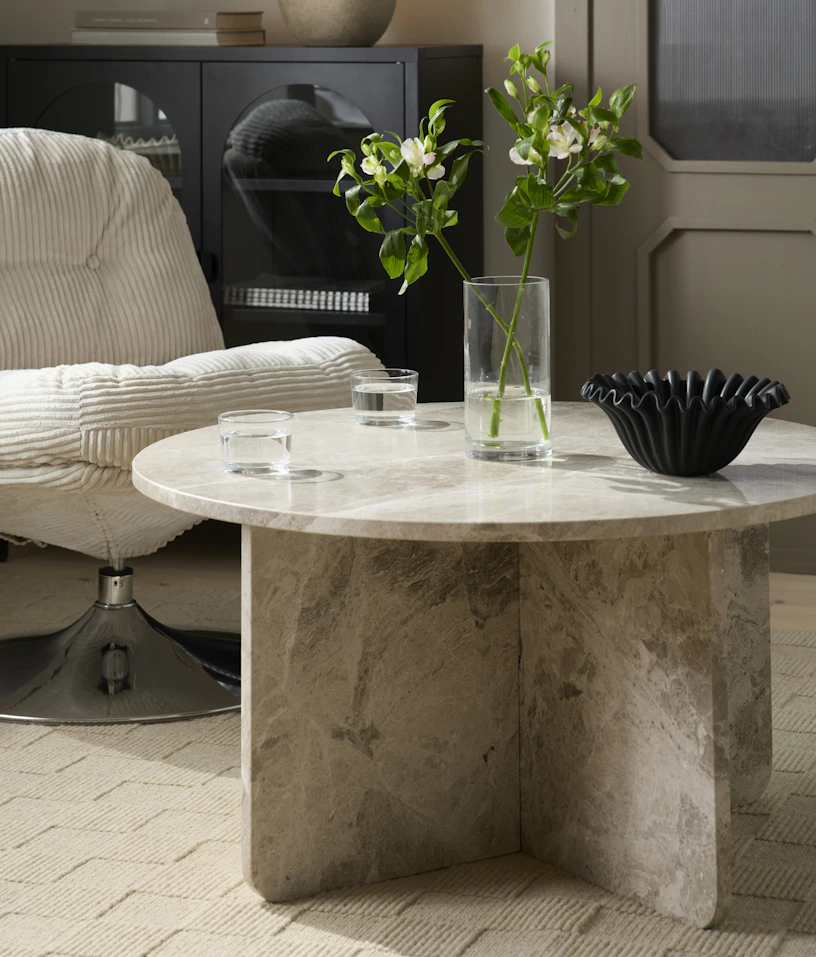 Ett runt soffbord i beige matmor står intill en fåtölj i beige manchester