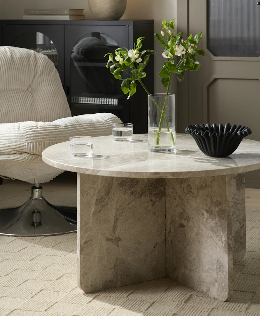 Ett runt soffbord i beige matmor står intill en fåtölj i beige manchester