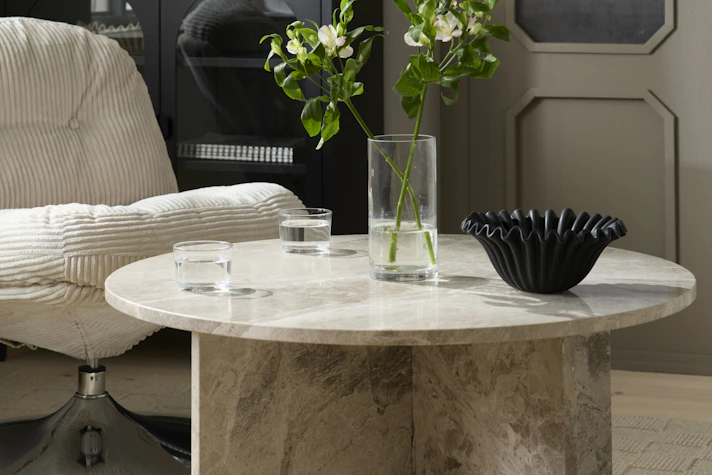 Ett runt soffbord i beige matmor står intill en fåtölj i beige manchester