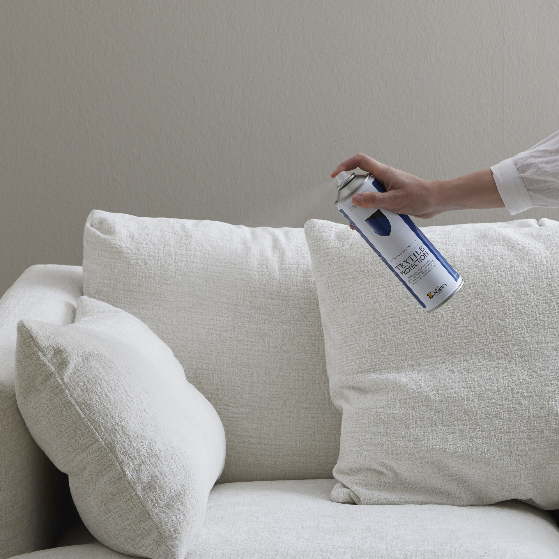 En hand håller fram en sprayburk med textilskydd framför en vit soffa