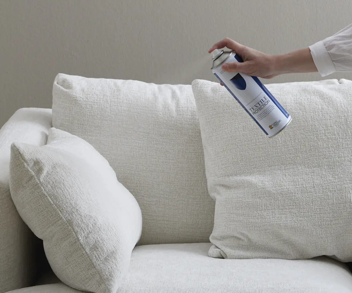 En hand håller fram en sprayburk med textilskydd framför en vit soffa