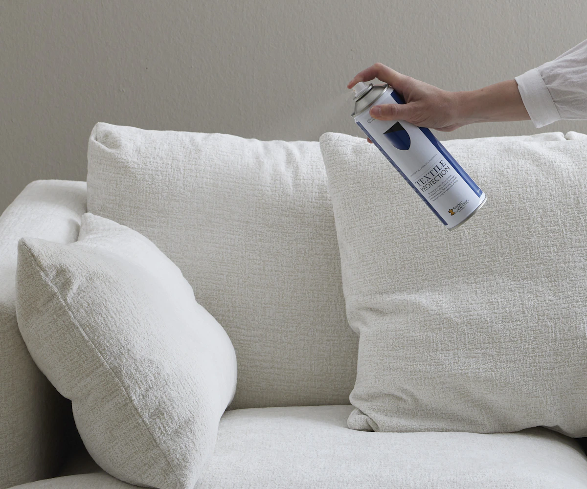 En hand håller fram en sprayburk med textilskydd framför en vit soffa