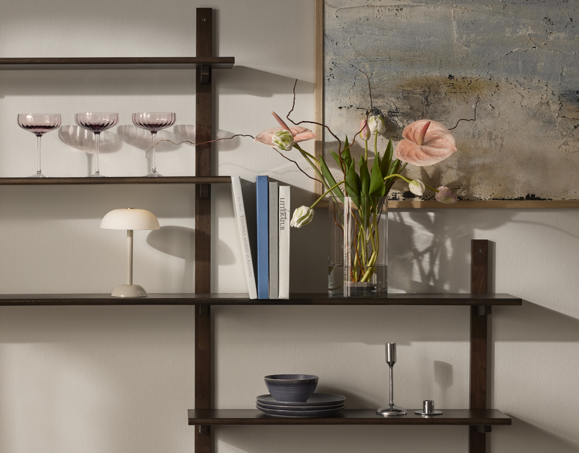 På hyllor i mörkt trä står en vas med vackra blommor i rosa toner, böcker en beige lampa och coupeglas