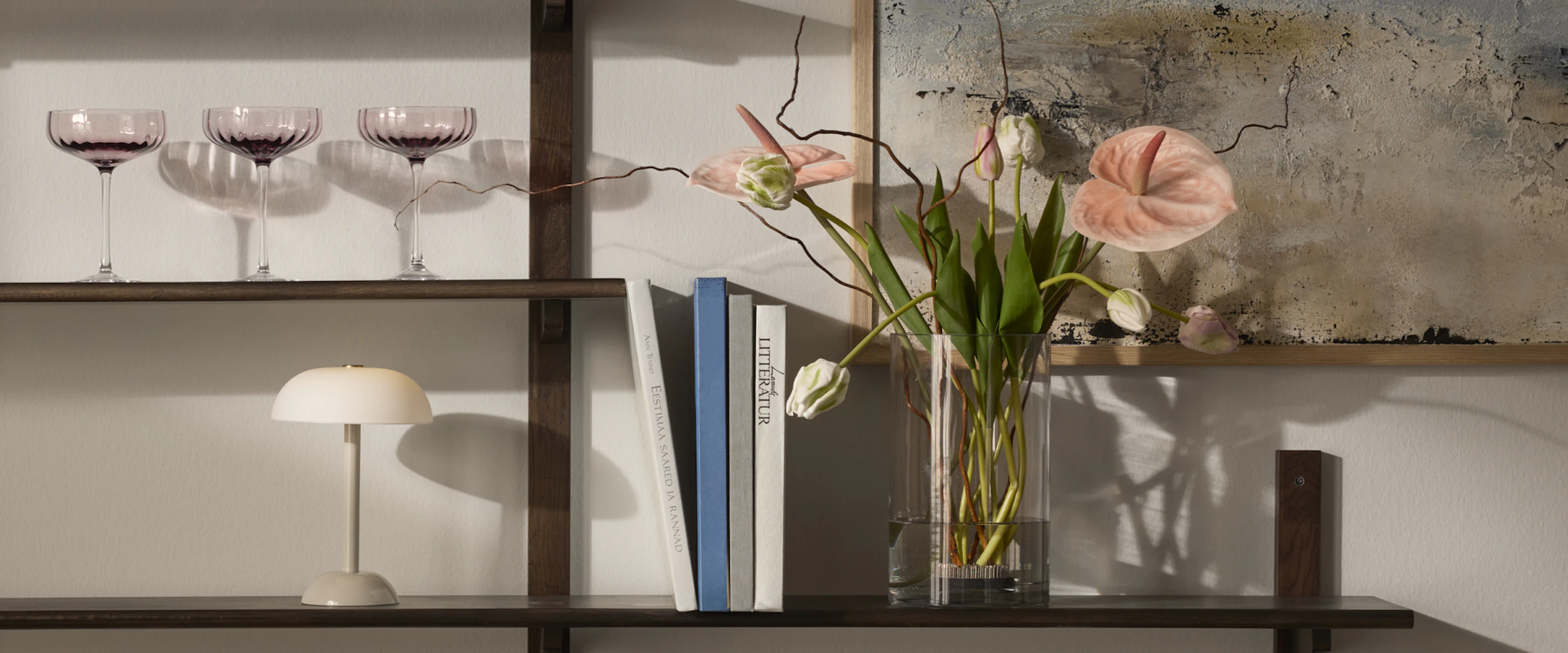 På hyllor i mörkt trä står en vas med vackra blommor i rosa toner, böcker en beige lampa och coupeglas