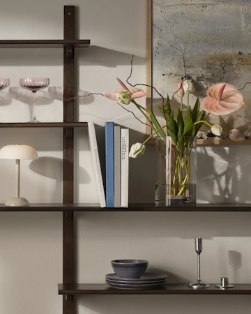 På hyllor i mörkt trä står en vas med vackra blommor i rosa toner, böcker en beige lampa och coupeglas