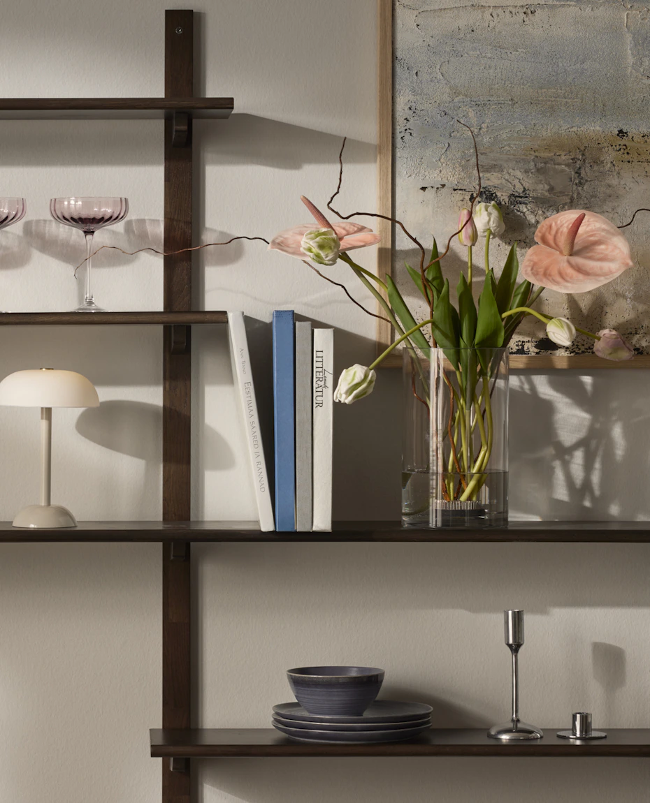 På hyllor i mörkt trä står en vas med vackra blommor i rosa toner, böcker en beige lampa och coupeglas