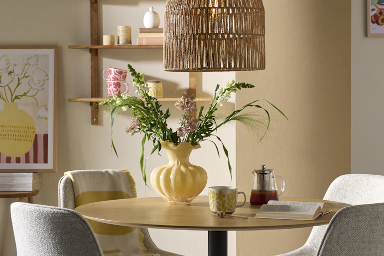 I ett ljust matrum står ett runt bord i ljust trä och fyra stolar i grått tyg. På bordet står en bukett i en gul vas.