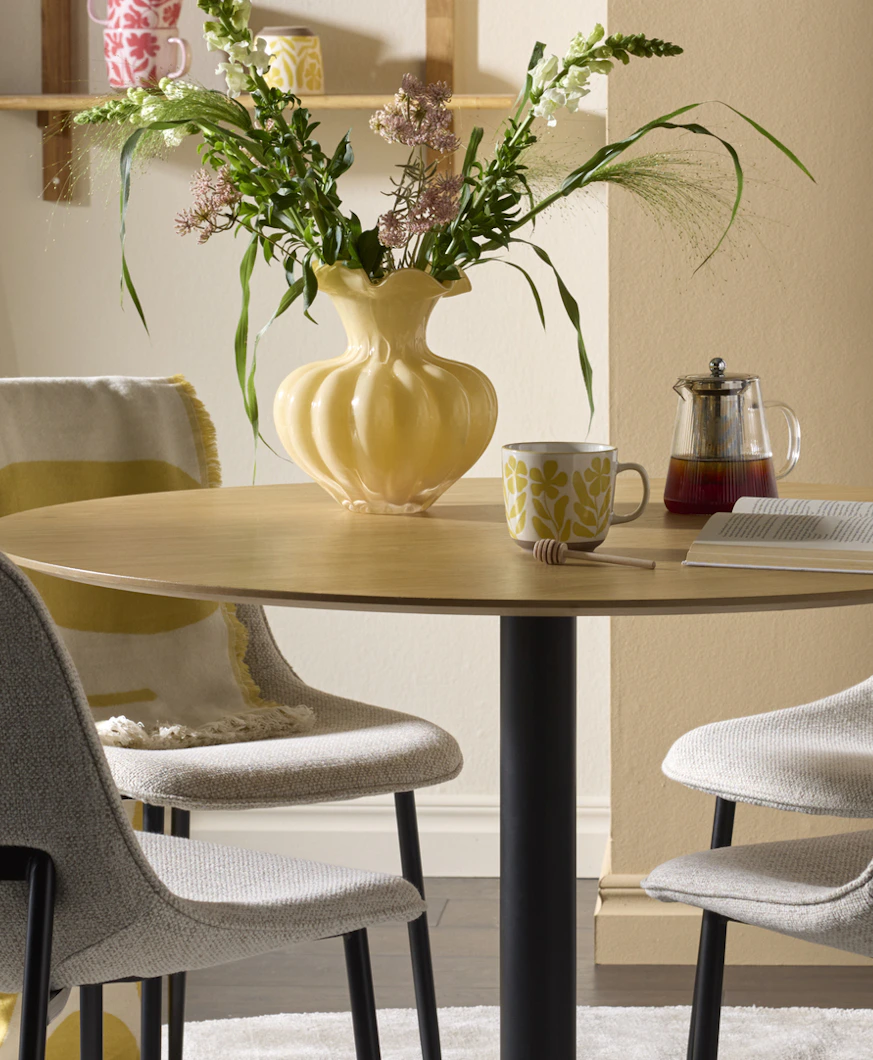 I ett ljust matrum står ett runt bord i ljust trä och fyra stolar i grått tyg. På bordet står en bukett i en gul vas.