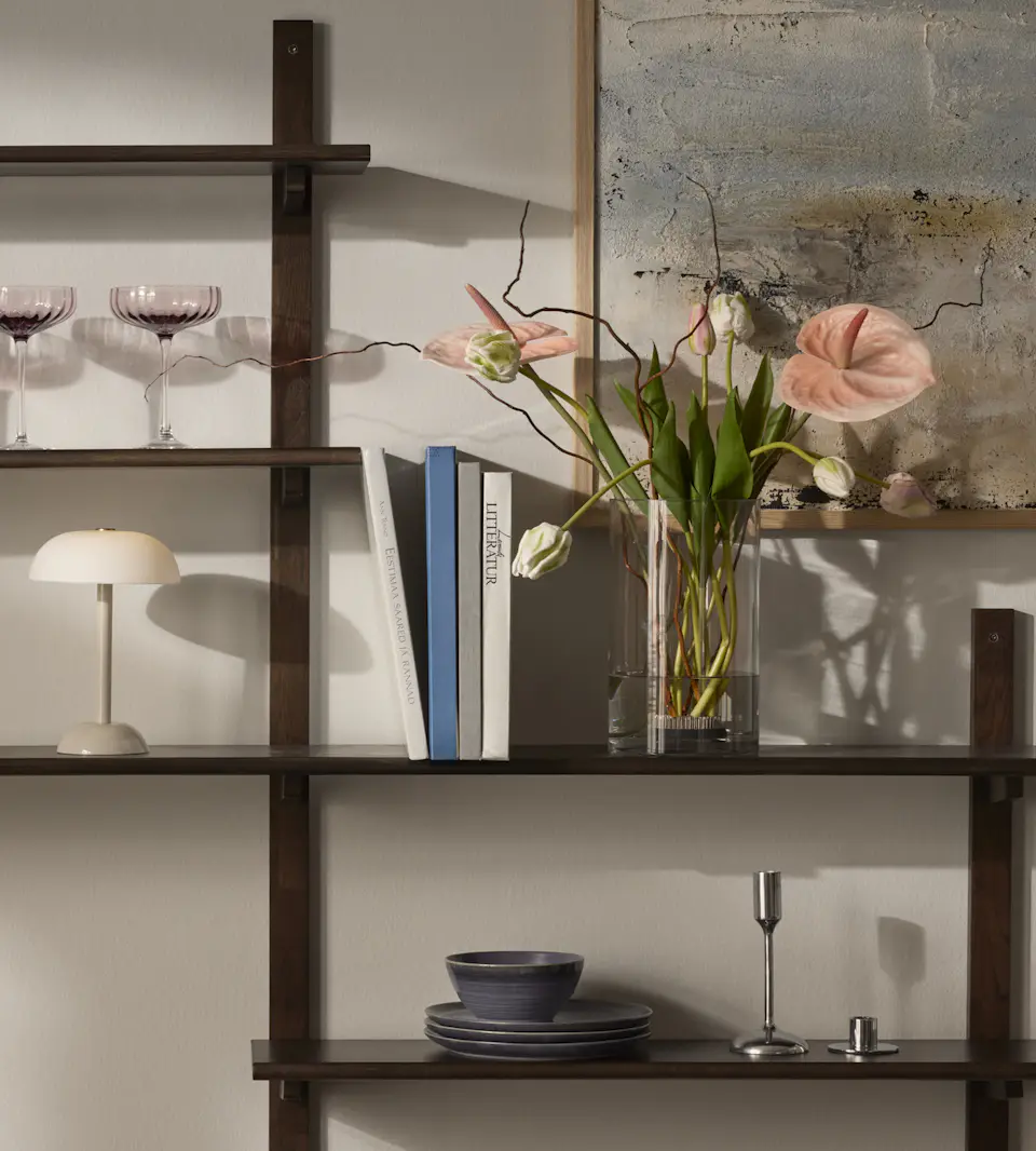 I en mörkbrun hylla står en vas med rosa snittblommor, en vit lampa och lite porslin. Bakom hyllan hänger en canvastavla med toner i blått och beige.