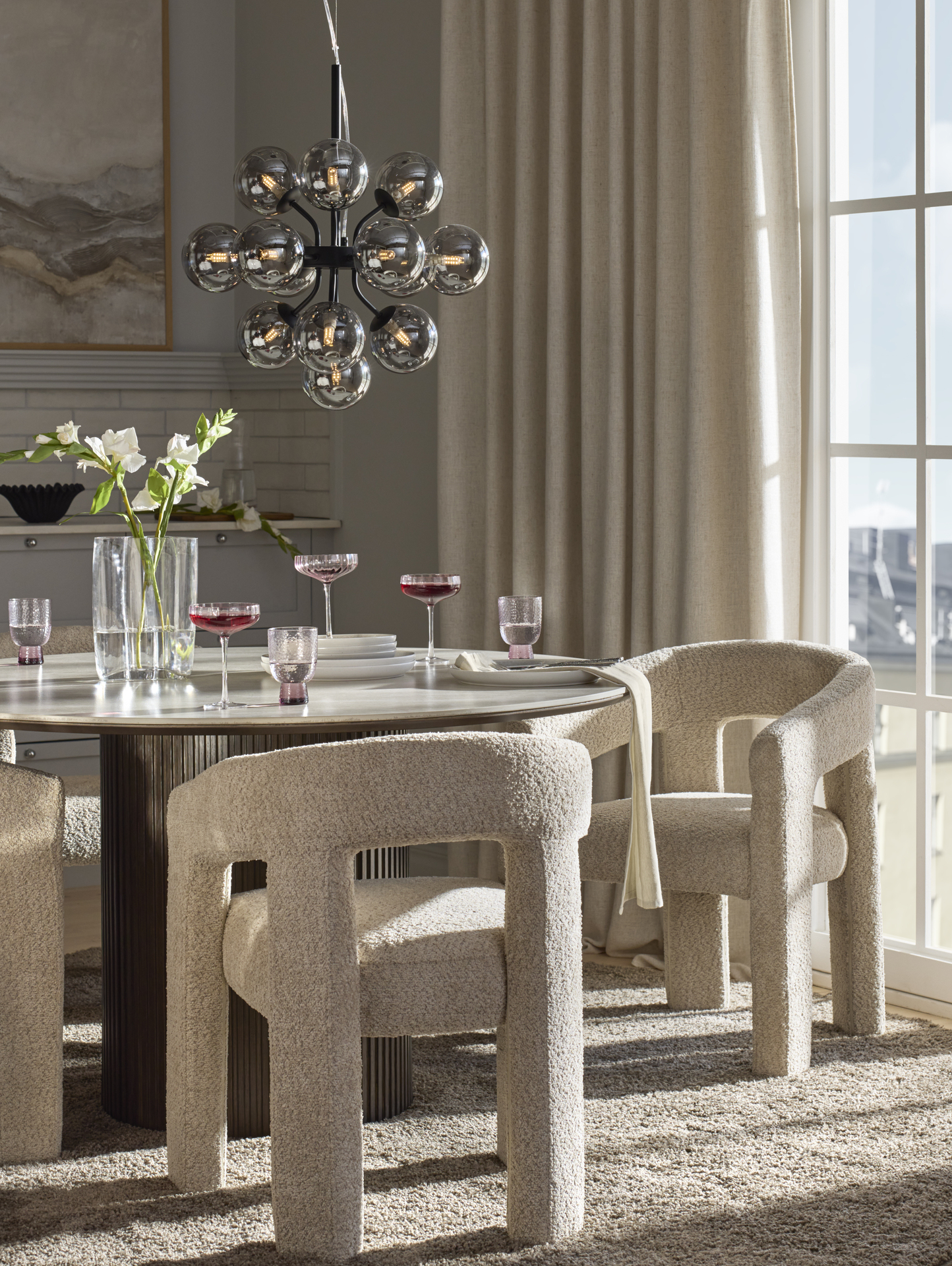 I ett grått kök står ett runt matbord med mörkbrun ribbad pelarfot och beigemelerad keramisk skiva. Runt bordet står karmstolar i beige bouclé. 