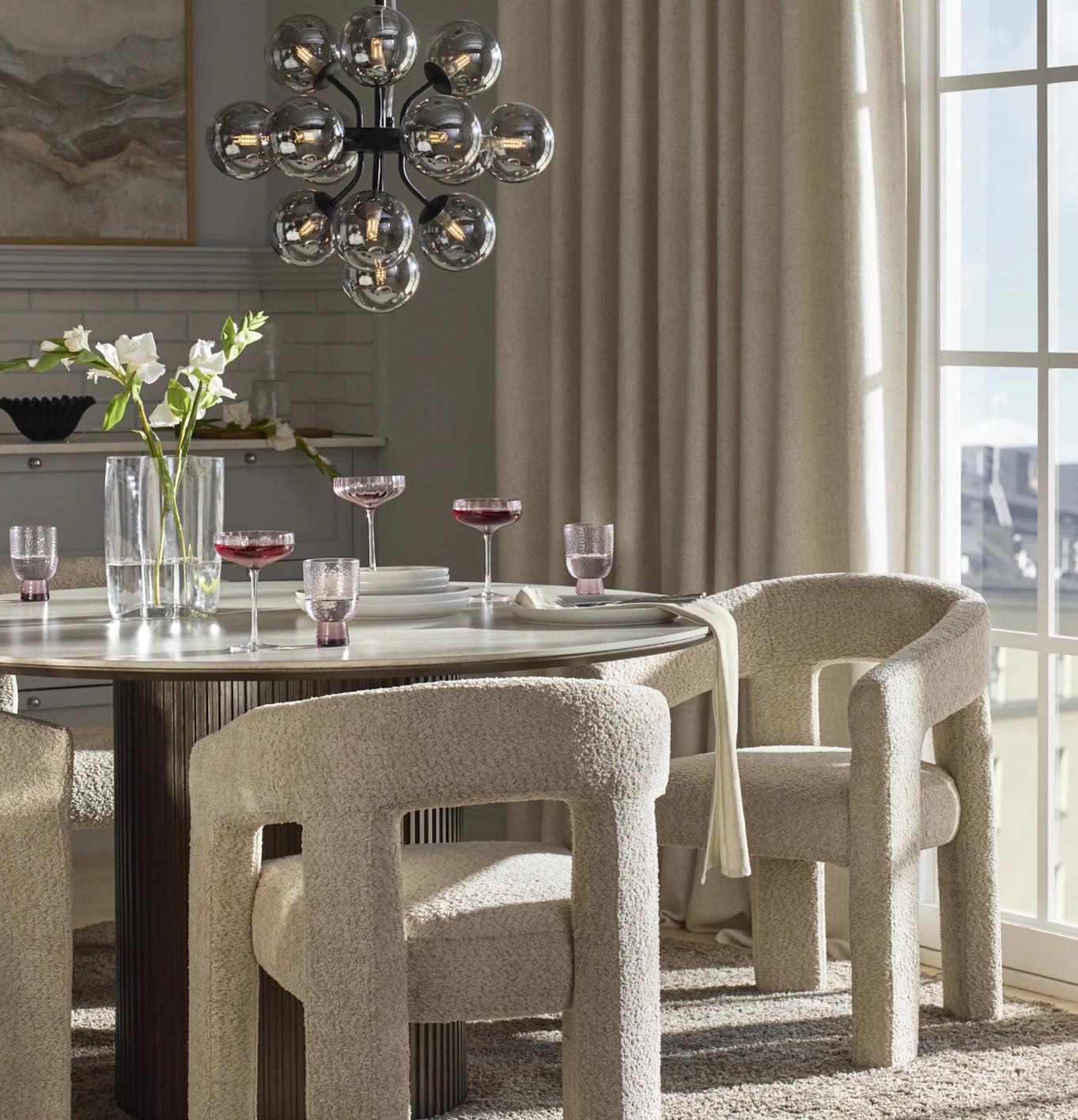 I ett grått kök står ett runt matbord med mörkbrun ribbad pelarfot och beigemelerad keramisk skiva. Runt bordet står karmstolar i beige bouclé.