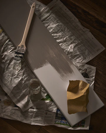 Ett hyllplan ligger på tidningspapper, målat till hälften i en varmgrå färg