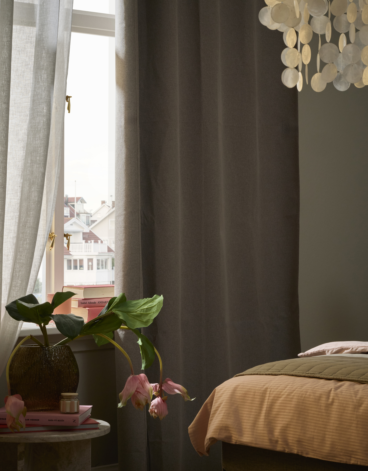 Vid fotändan av en säng står ett sidobörd med en vas framför ett fönster med mörkläggande gardiner