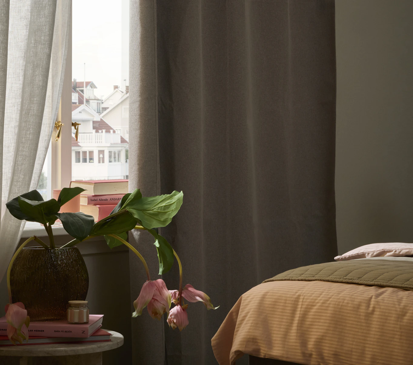 Vid fotändan av en säng står ett sidobörd med en vas framför ett fönster med mörkläggande gardiner