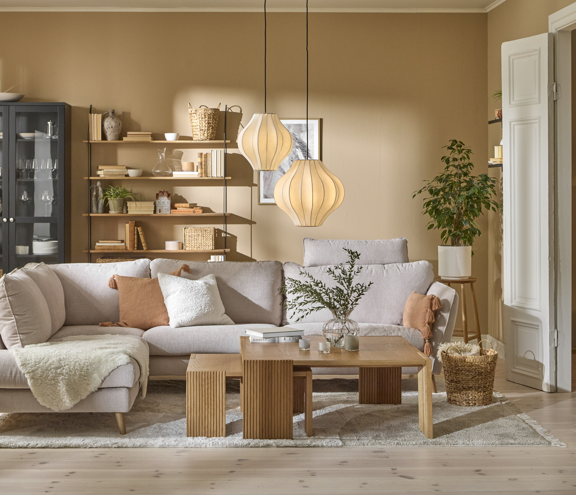 I ett gult vardagsrum står en ljusgrå soffa framför ett svart vitrinskåp och en vägghylla med hyllplan i trä